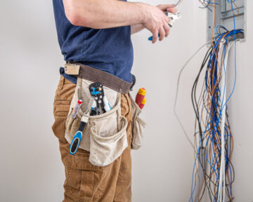 Electrician builder at work, examines the cable connection in the electrical line in the fuselage of an industrial switchboard. Professional in overalls with an electrician's tool.